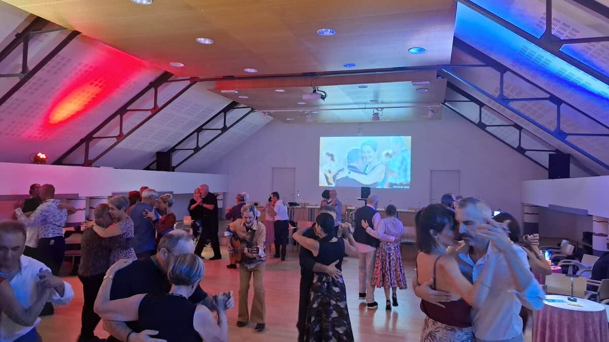 Des couples dansent ensemble dans une salle chaleureuse éclairée de lumières colorées, créant une ambiance conviviale et détendue.