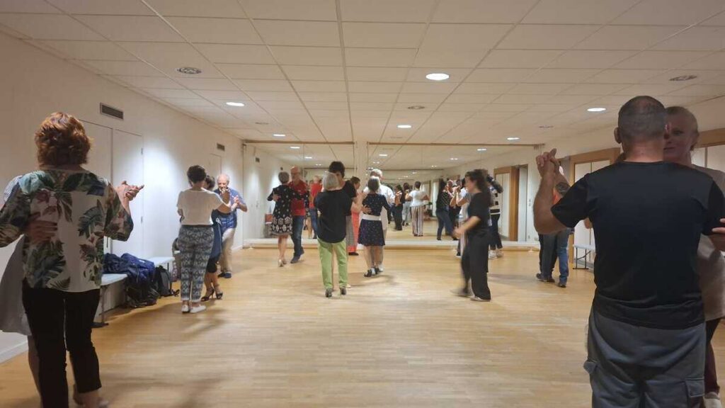 Un groupe de personnes pratique une danse en salle, devant un grand miroir, dans une ambiance détendue et conviviale.