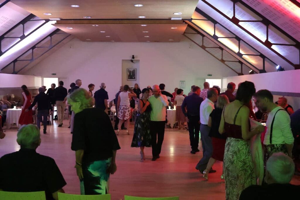 Une salle animée où de nombreuses personnes discutent et dansent, dans une ambiance chaleureuse avec des éclairages colorés qui donnent une atmosphère festive.